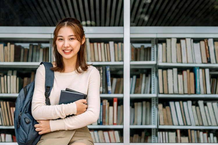 Student with backpack holding a book, sitting in front of a bookshelf at a library.